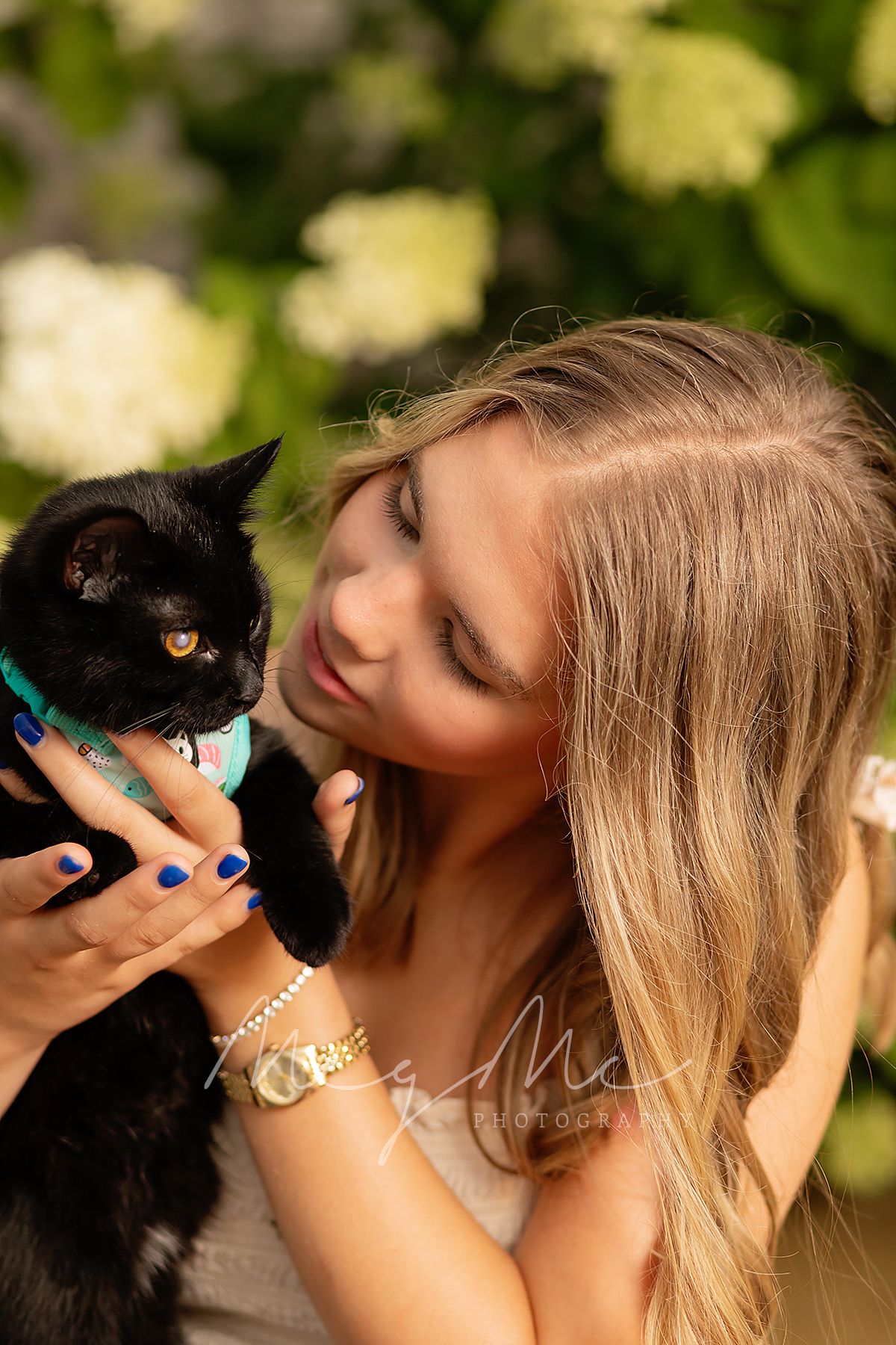 Black cat wearing turquoise bandana held outdoors against greenery and golden sunlight.