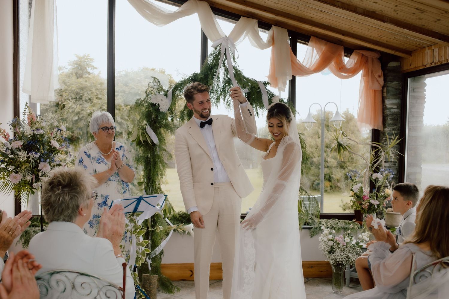 A wedding ceremony takes place on a sunlit porch decorated with plants and white curtains.
