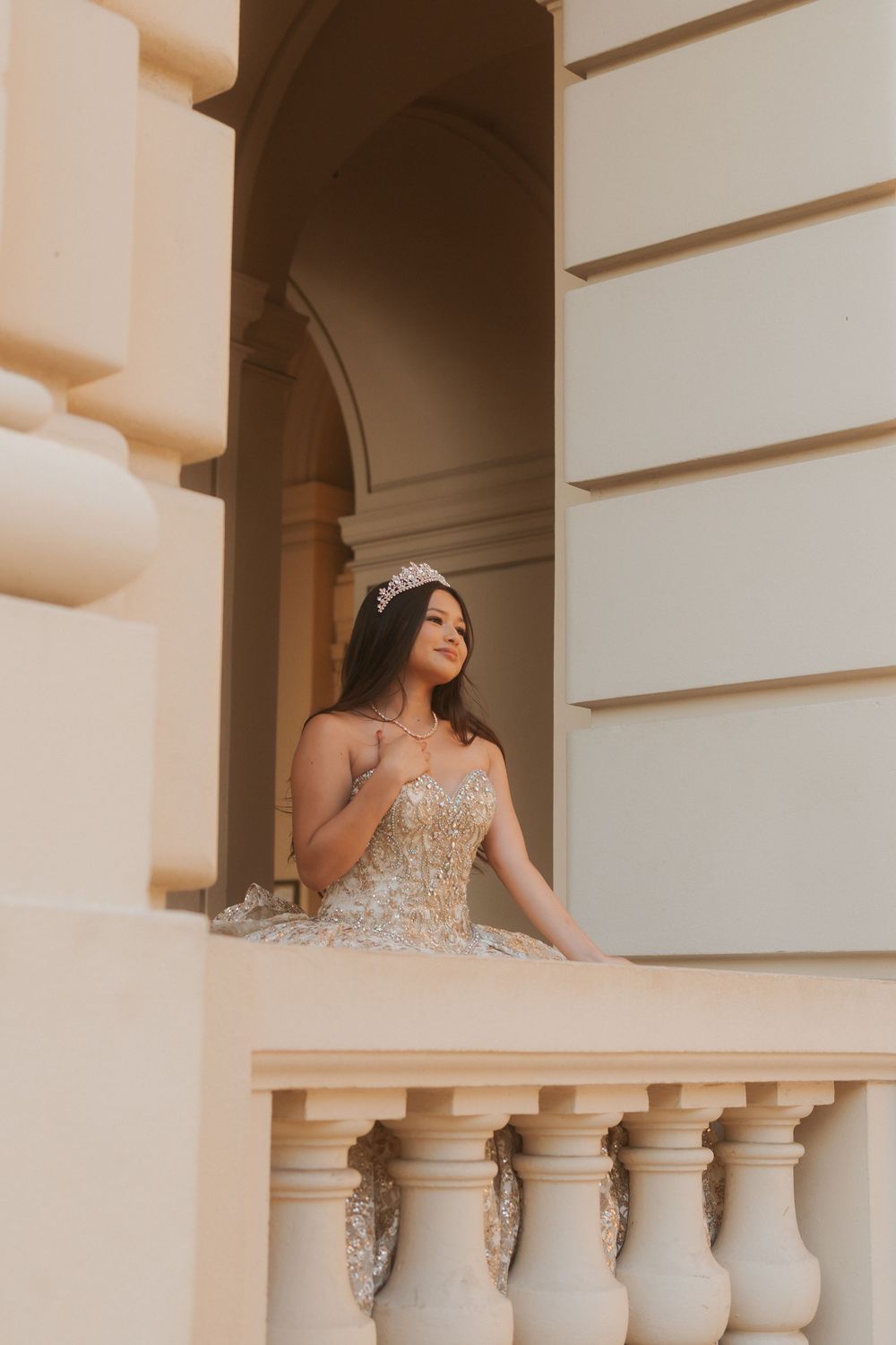 A person in an elegant beaded gown poses on a balcony with white columns in a dreamy sepia-toned setting.