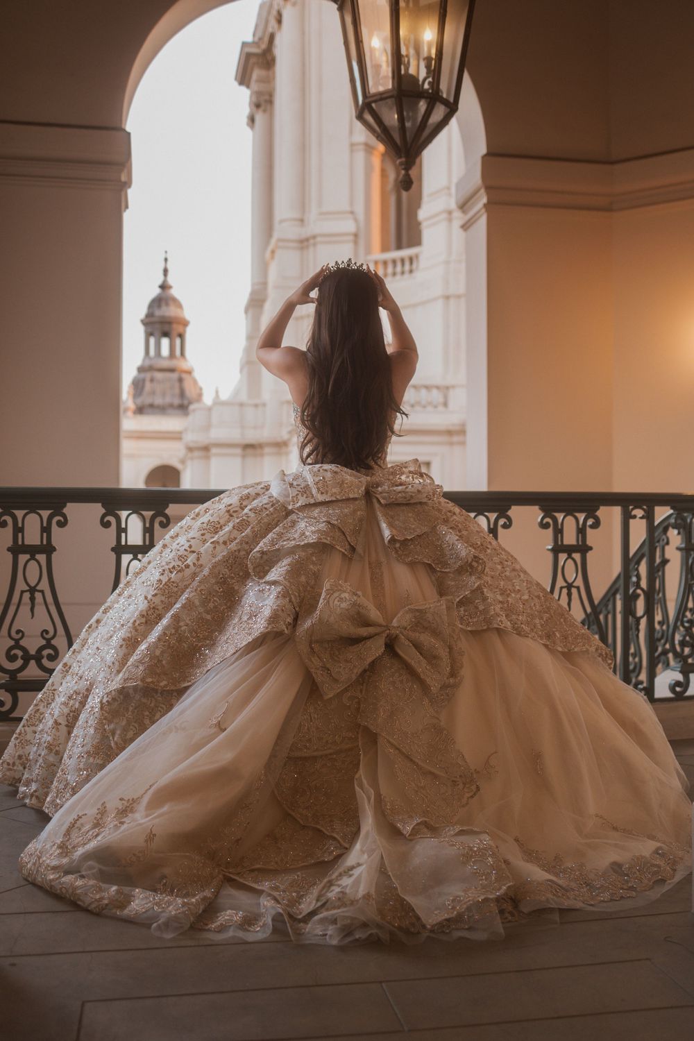 A person in an ornate white ball gown stands on a balcony overlooking a cathedral tower at sunset.