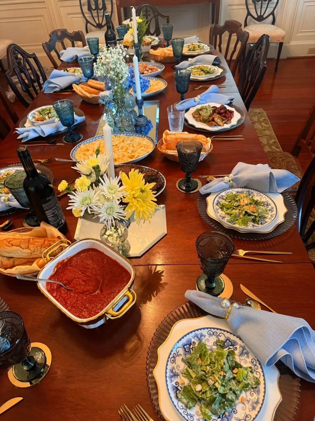 A formal dinner table set with salad, pasta, red sauce, cheese plates and yellow flower centerpiece on dark wooden table.