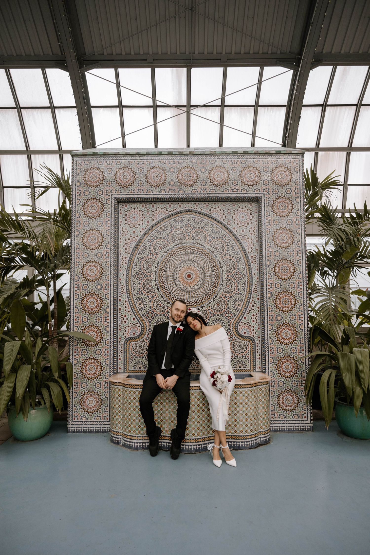 A couple sits together against an ornate Moroccan-style tile wall backdrop in a greenhouse with potted palm plants.
