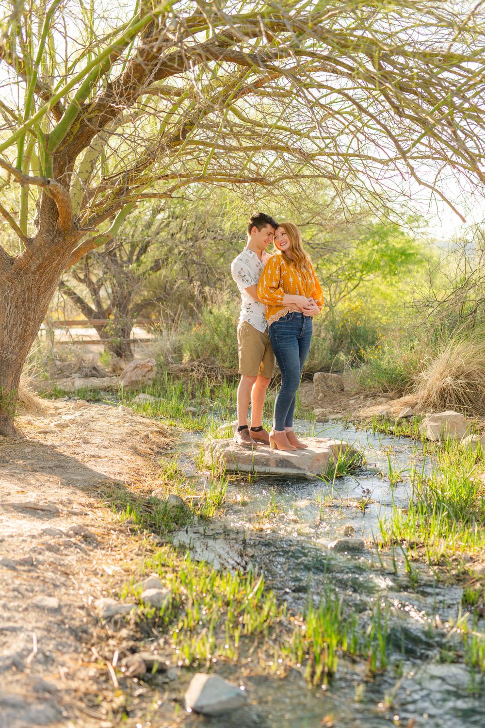 A romantic scene by a stream with a couple standing on rocks under a desert willow tree.