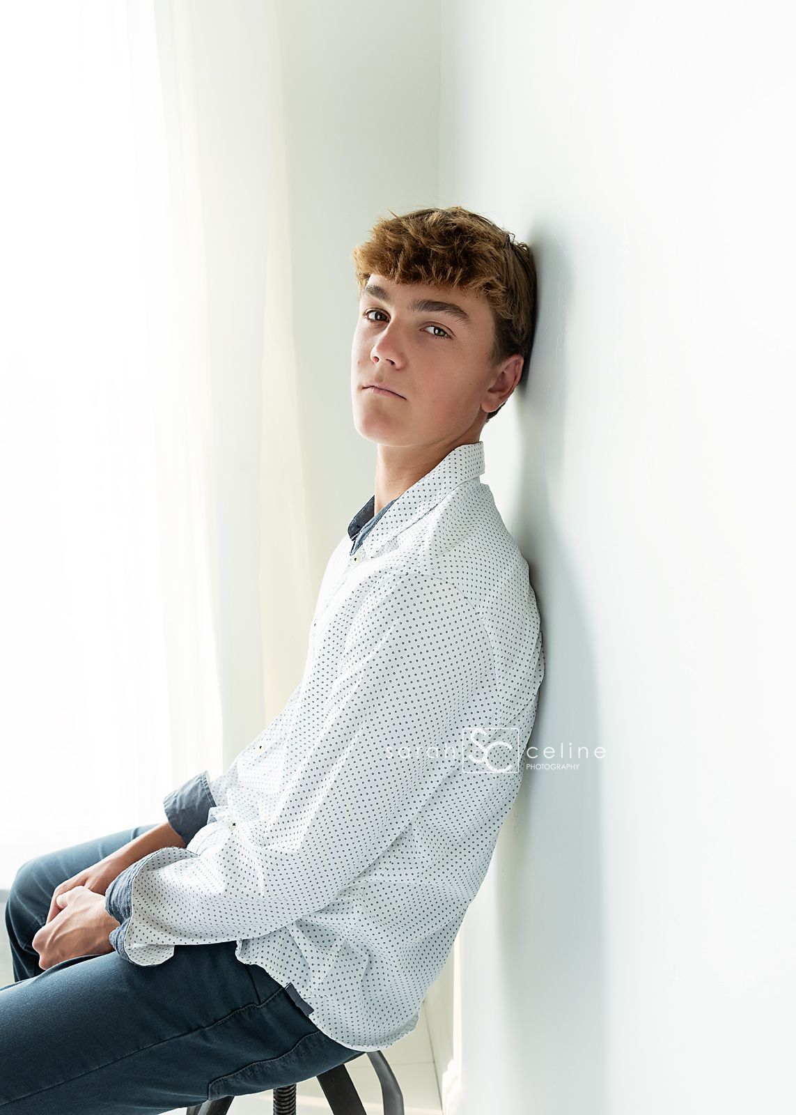 A young person poses in a white polka dot dress shirt and jeans against a light-colored wall in a studio setting.