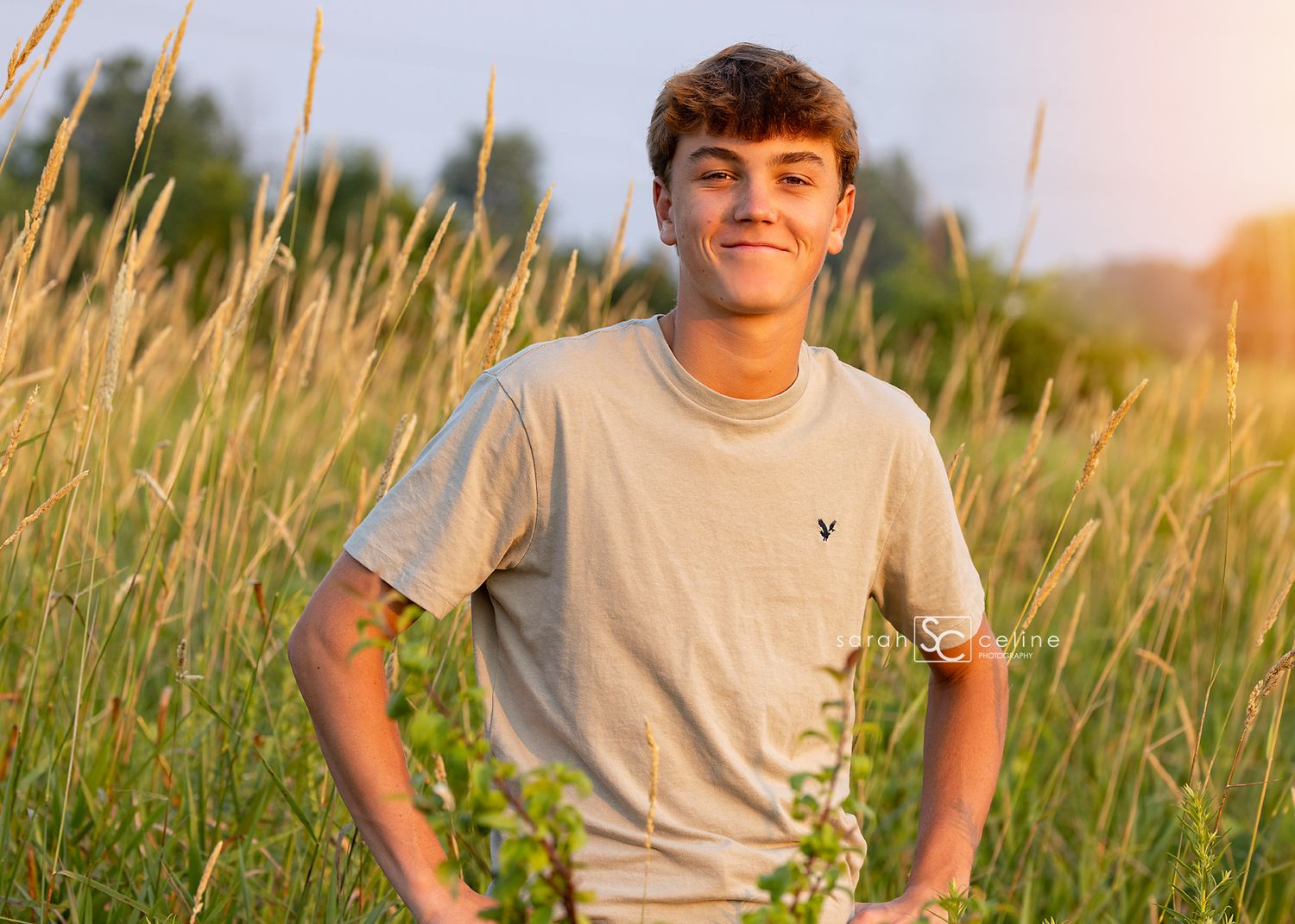 A young person in a beige t-shirt stands smiling in a sunlit field of tall grass during golden hour.
