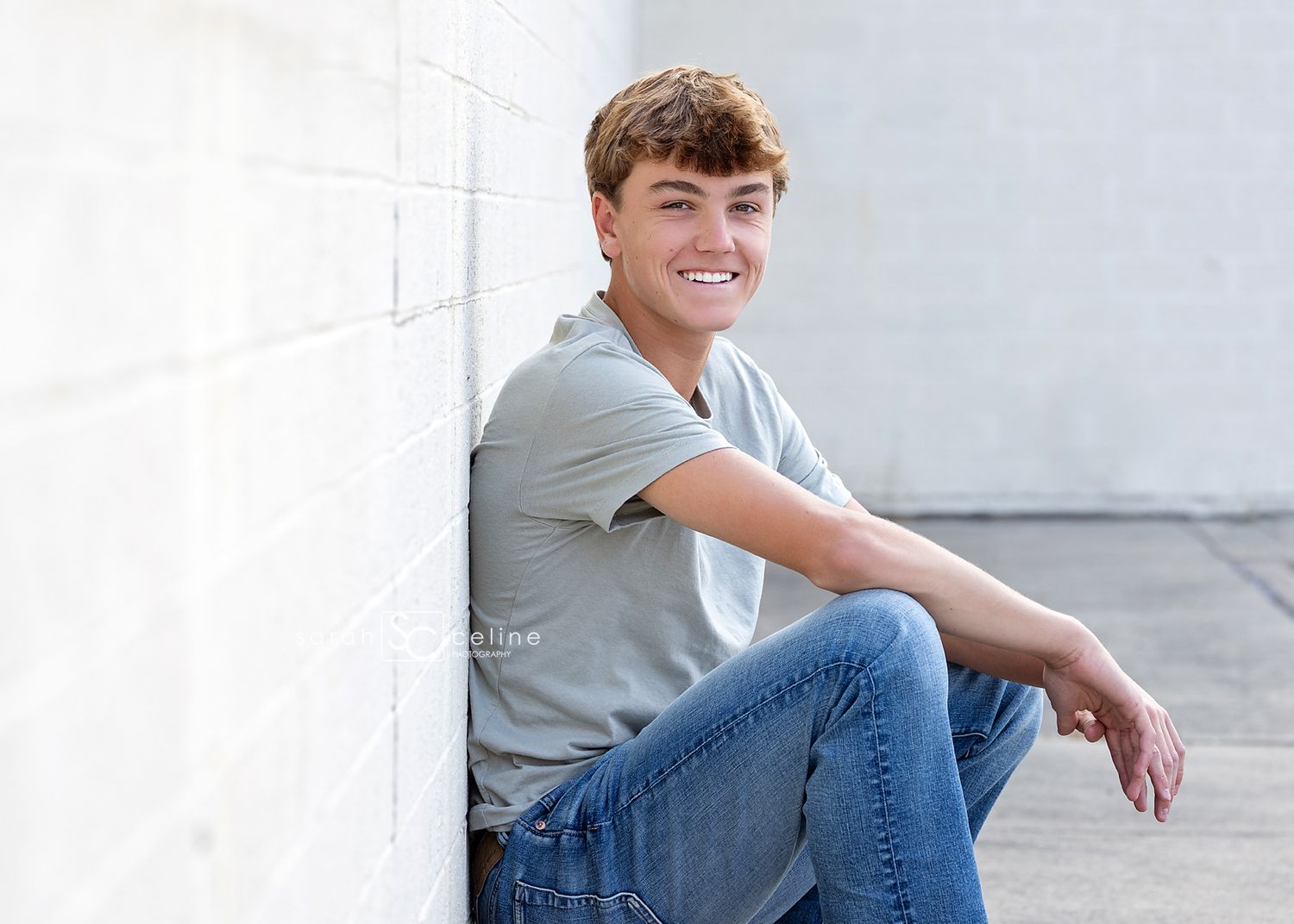 Young person in blue jeans and gray shirt sitting casually outdoors for a portrait session against light background.