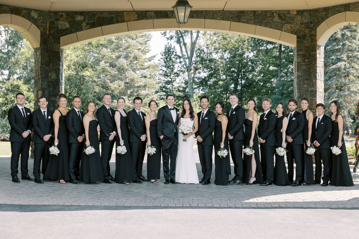 A formal wedding party poses together outdoors under a covered stone portico during a summer celebration.
