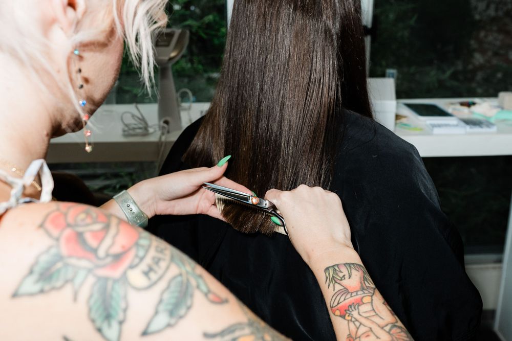 A hairstylist with tattooed arms cuts long dark hair at a salon workstation.