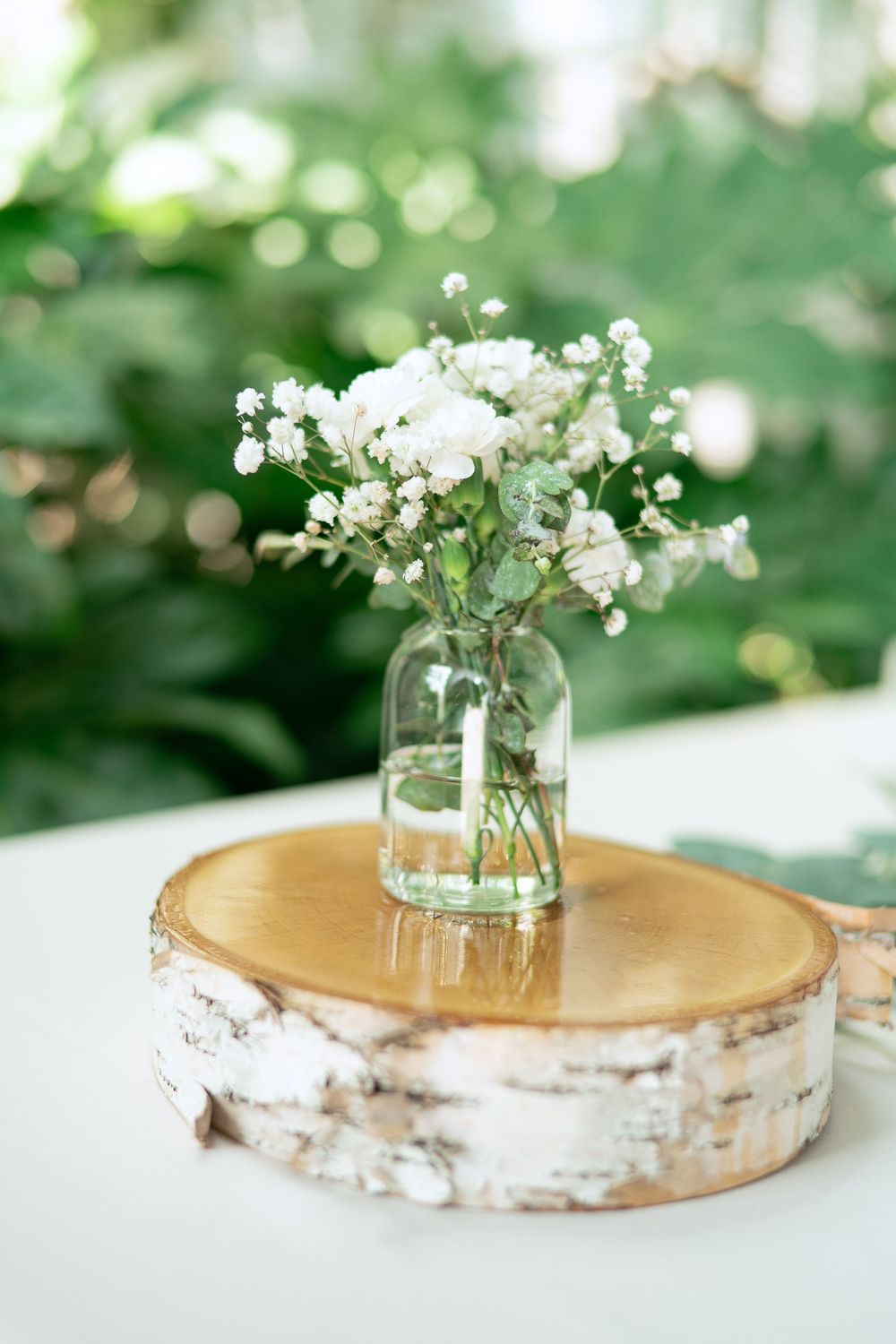A delicate bouquet of white baby's breath flowers in a glass jar sits on a natural birch wood slice centerpiece.