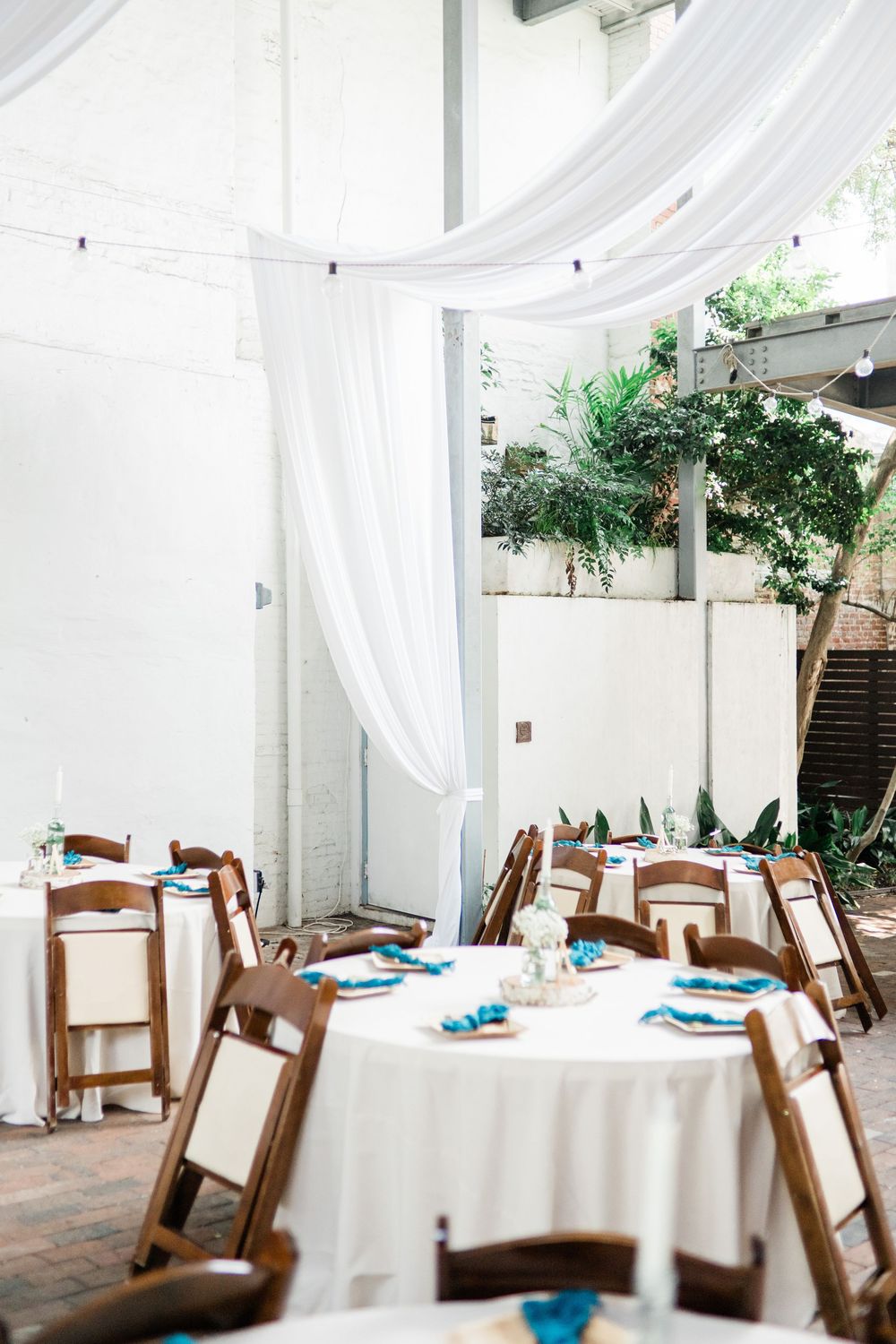 Round tables with white tablecloths and wooden chairs set beneath elegant white draping and string lights in an airy venue.