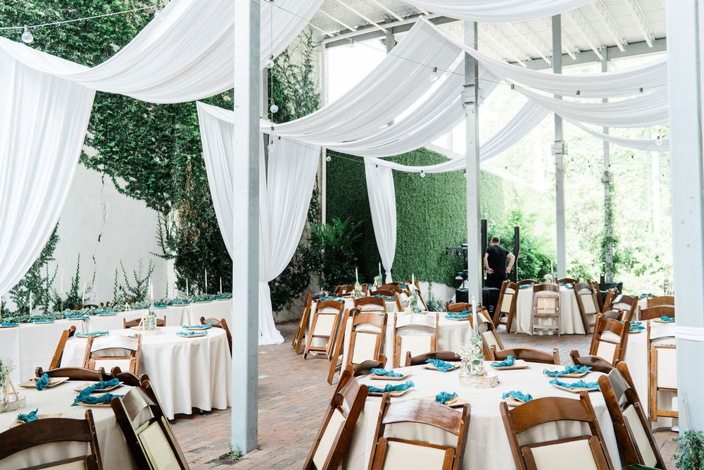 Elegant outdoor wedding reception setup with white draping canopy, rustic wooden chairs and round tables decorated in blue.