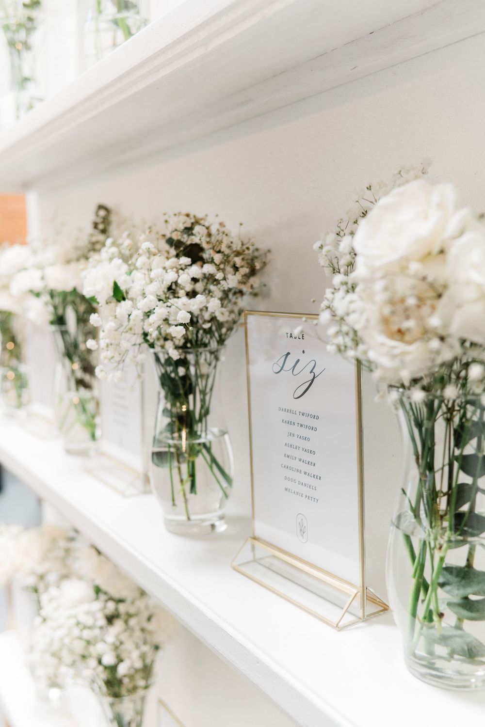 White baby's breath and floral arrangements in glass vases line a white shelf with a framed wedding menu display card.