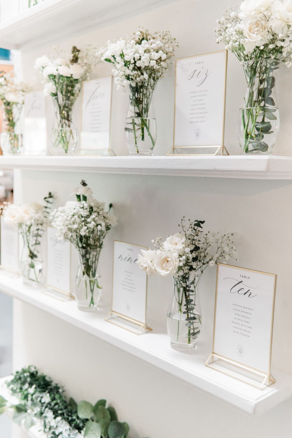 Wedding seating chart display with elegant white floral arrangements and framed table assignments on white shelves.