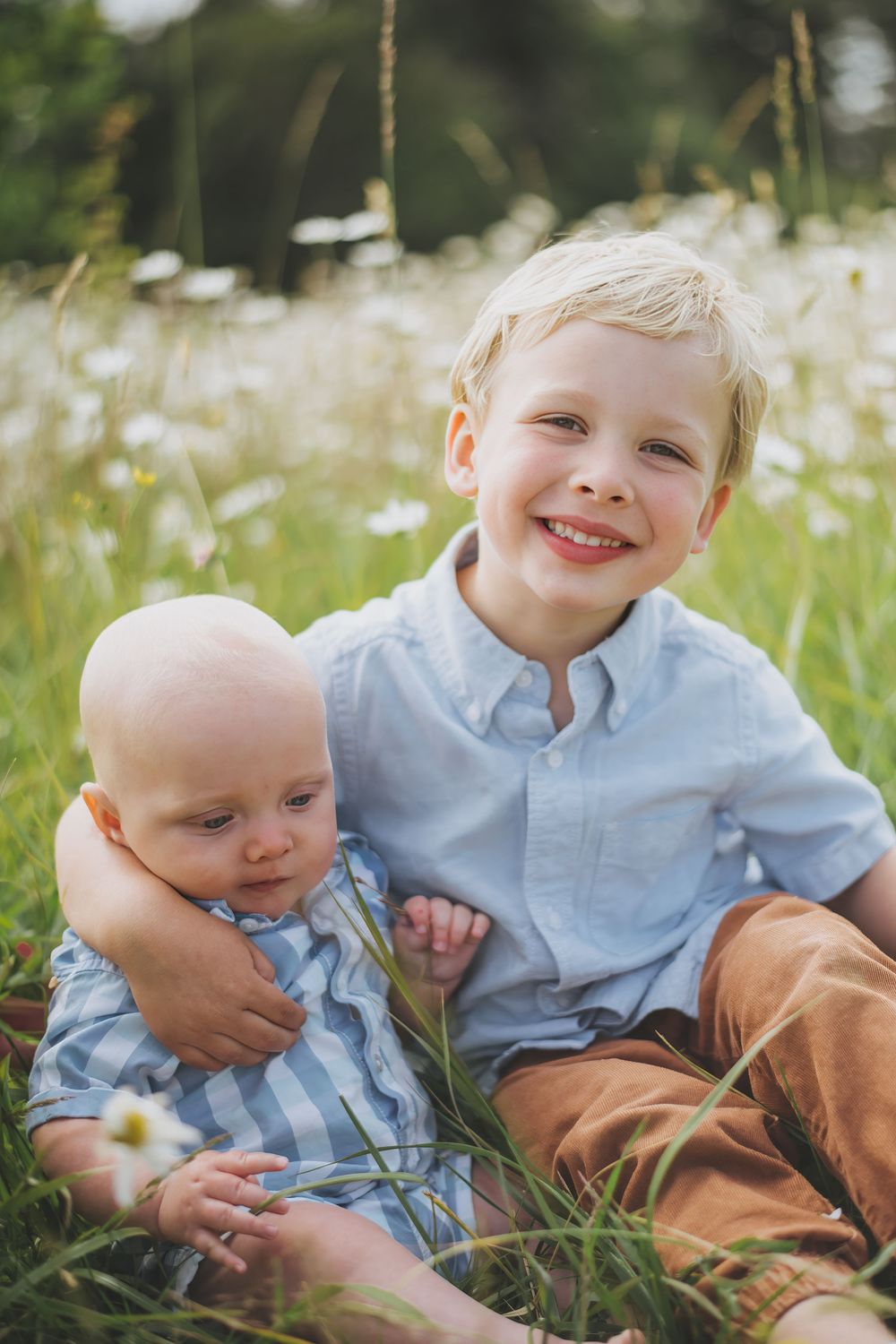 Two children wearing blue shirts sit together in a field of white daisies and grass during summer.