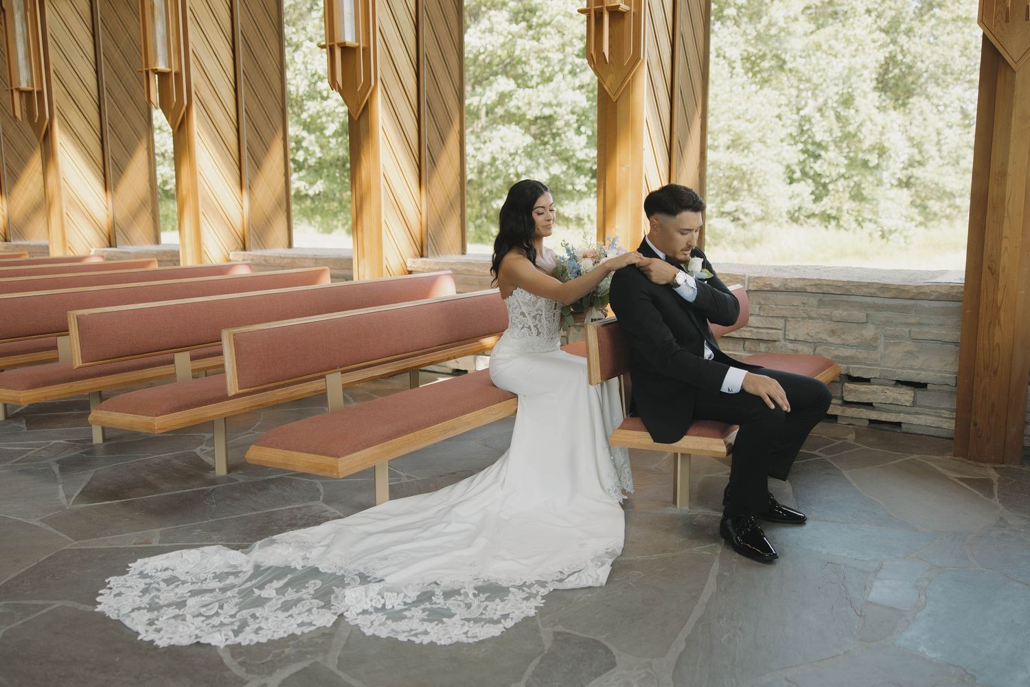 Two people in wedding attire sit together on a wooden bench in an outdoor chapel with dramatic shadows cast from above.