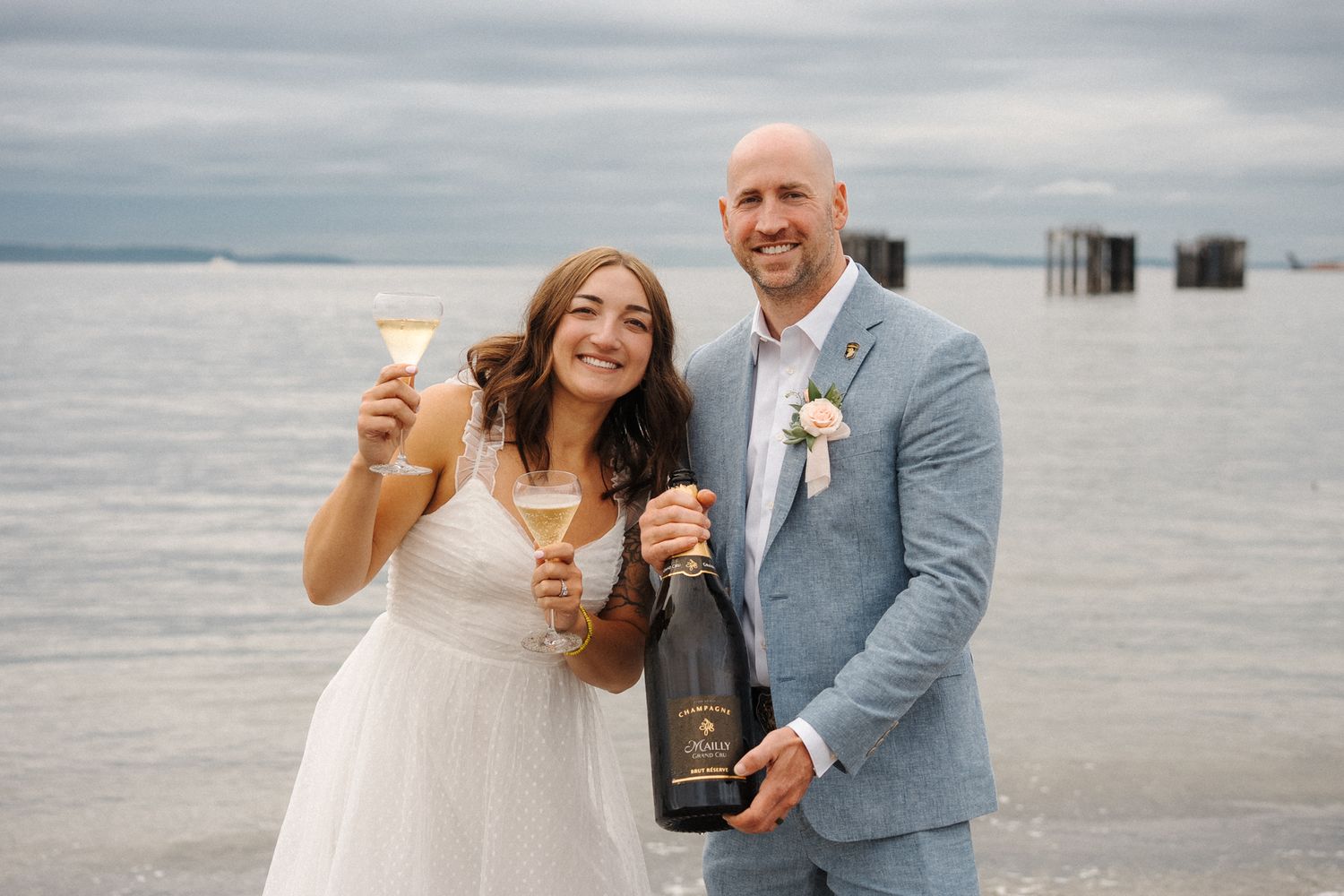 A couple shares a celebratory moment with champagne glasses and a bottle at a beachside location.