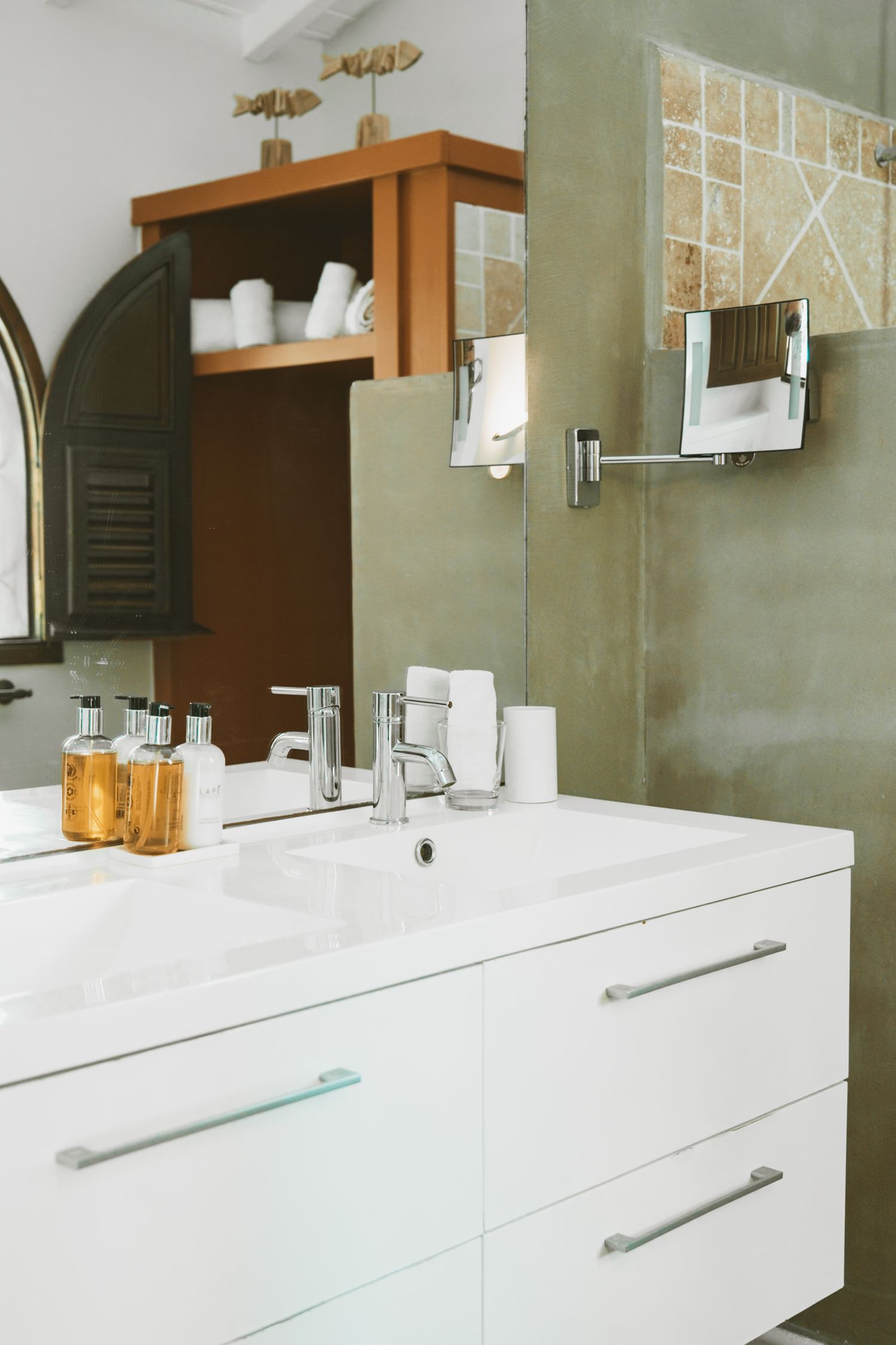 Modern white bathroom vanity with sage green walls, wooden shelving, and amber glass bottles on the counter.