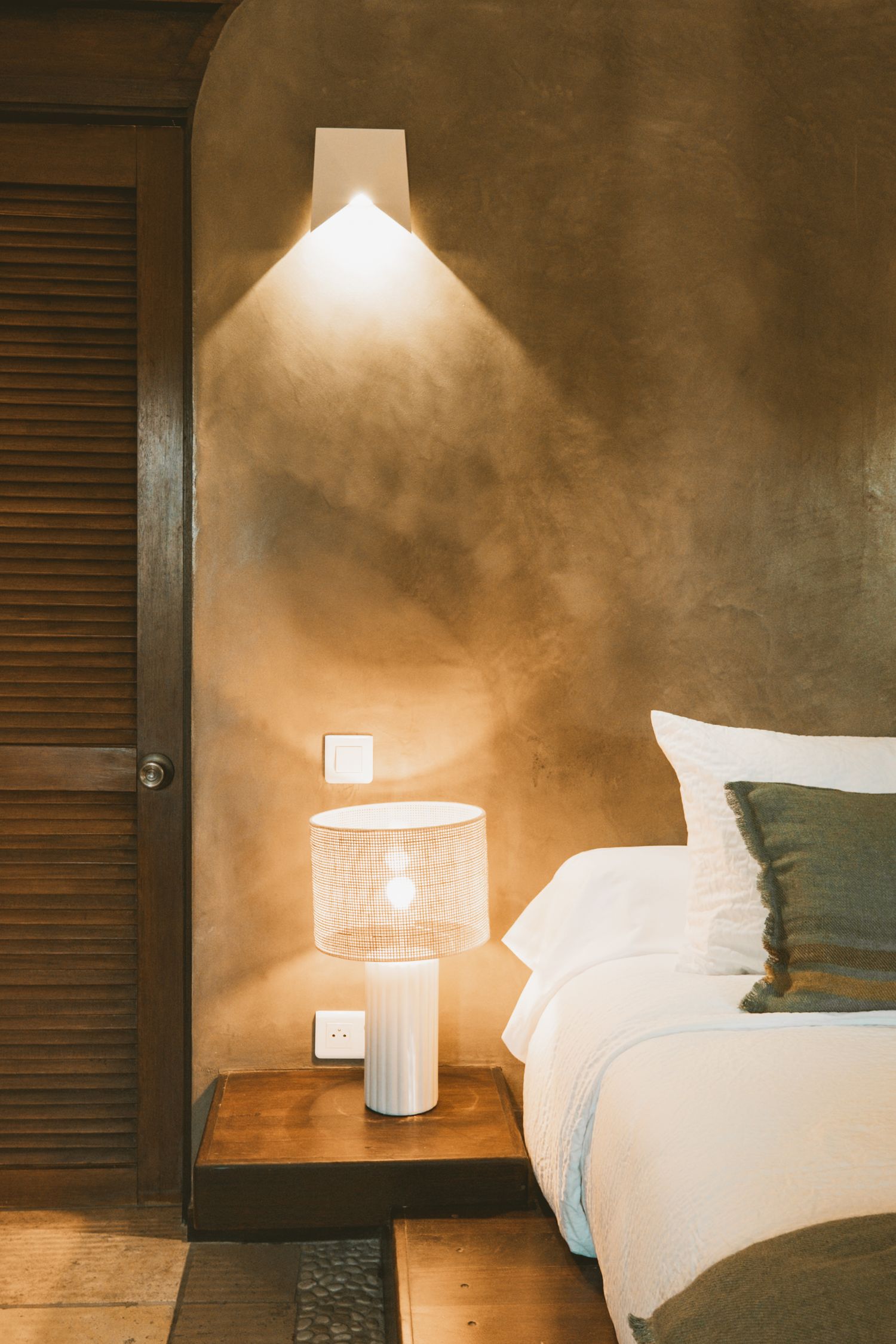 Cozy bedroom interior with warm lighting from wall sconce and table lamp illuminating white bedding and wooden furniture.