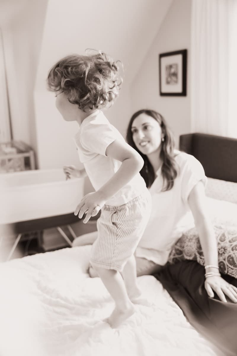 A toddler stands on a bed in striped shorts while being watched from behind in a black and white photograph at family Chicagoland session