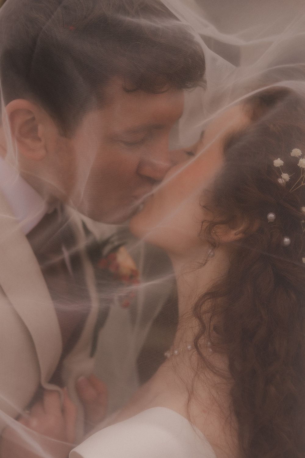 A bride and groom share a tender kiss beneath a flowing wedding veil, captured in a soft, romantic close-up portrait.