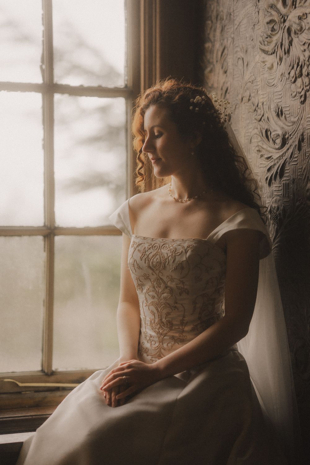 A bride in a lace gown sits by a sunlit window, gazing downward with a serene expression.