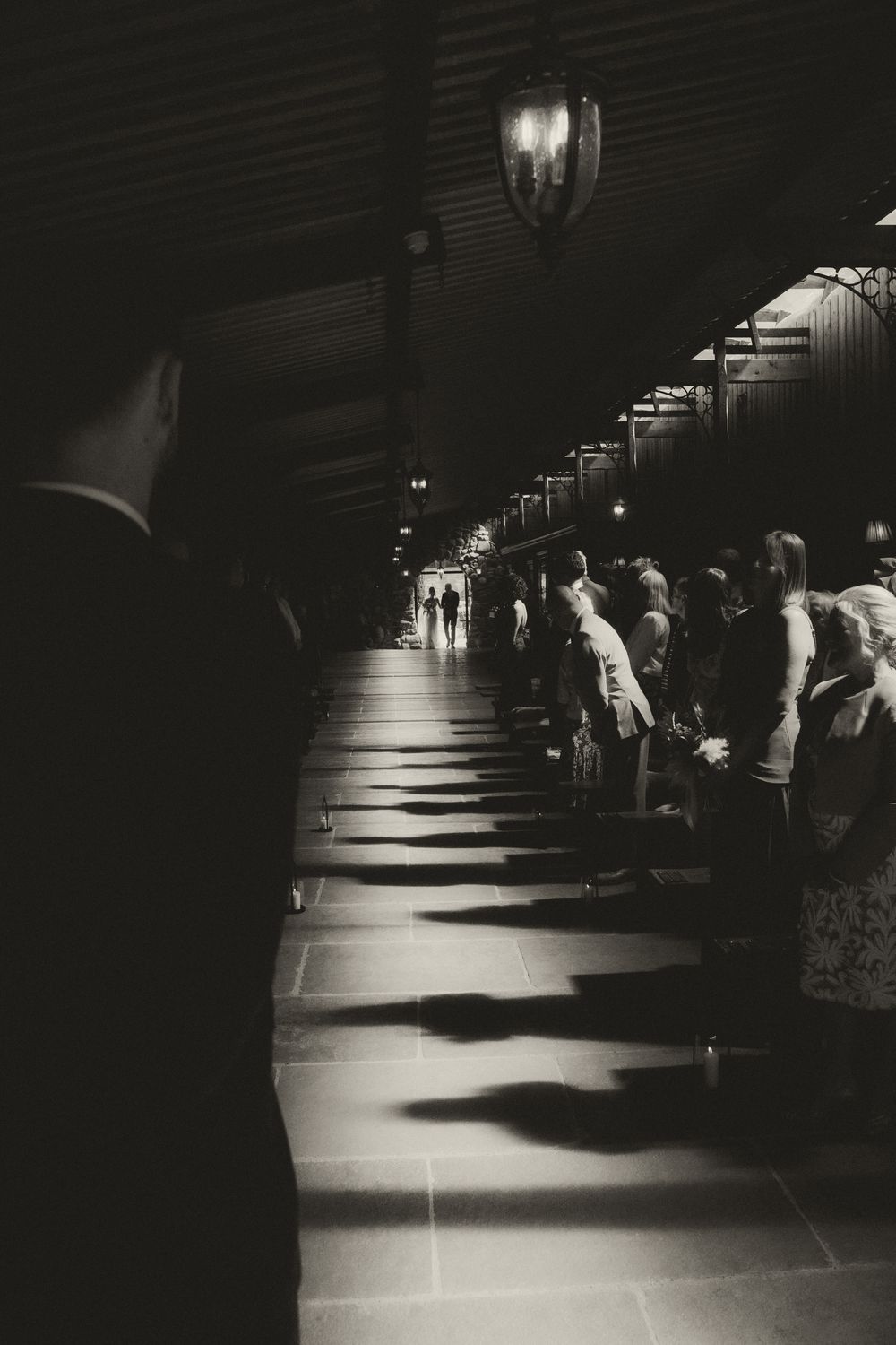 Black and white photo of elegantly dressed guests lining a dimly lit corridor with dramatic shadows and hanging lanterns.