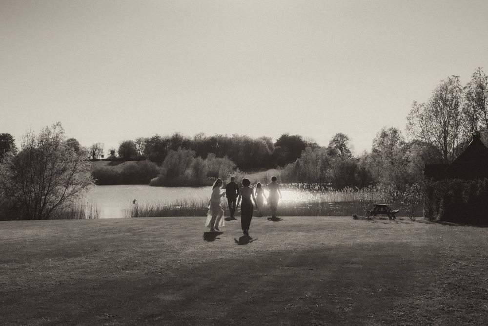 Black and white photo of people standing on a grassy field near a tranquil lake surrounded by trees.