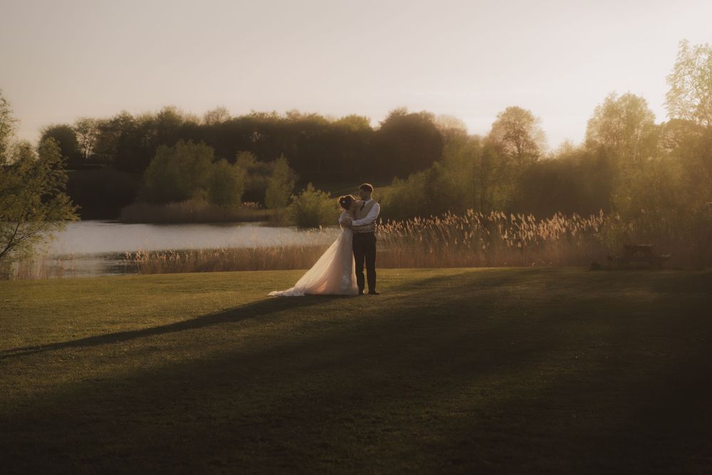 Couple embracing at golden hour sunset by a lake, bride in flowing white gown with long train on lush green lawn.