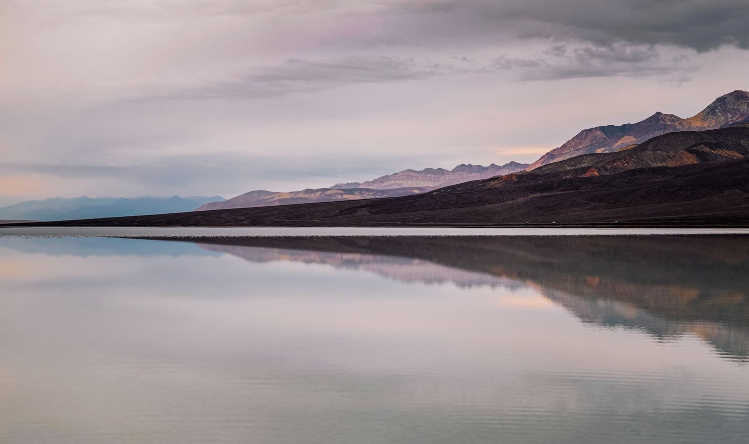 Serene mountain reflection on still lake water under soft pastel cloudy sky at dusk.