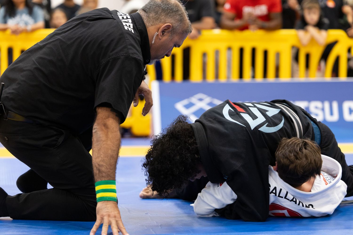Martial artists in black gis competing in a grappling match on a blue mat.