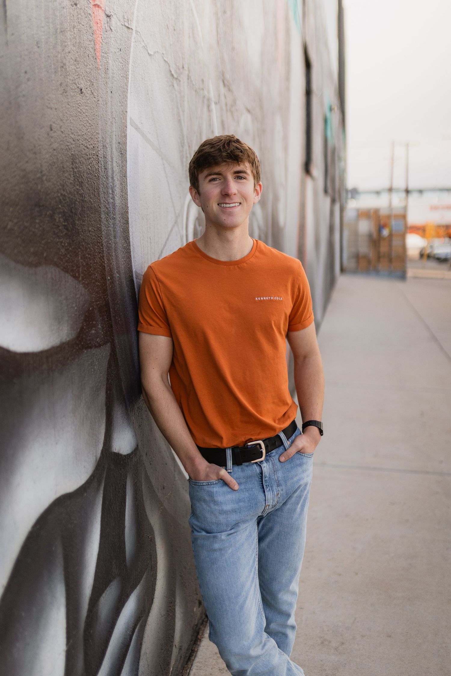 Person in orange t-shirt and light jeans poses confidently against concrete wall in urban setting.