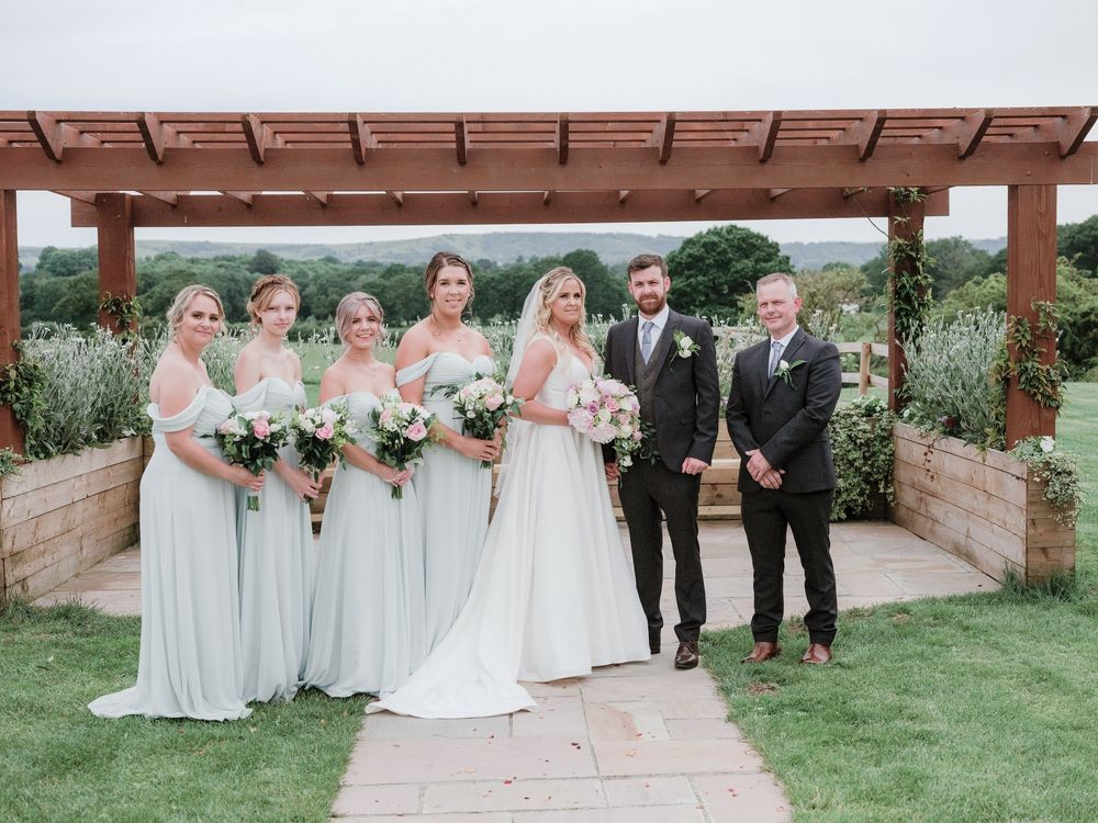 A wedding party poses under a wooden pergola in a scenic garden setting wearing mint green and black formal attire.