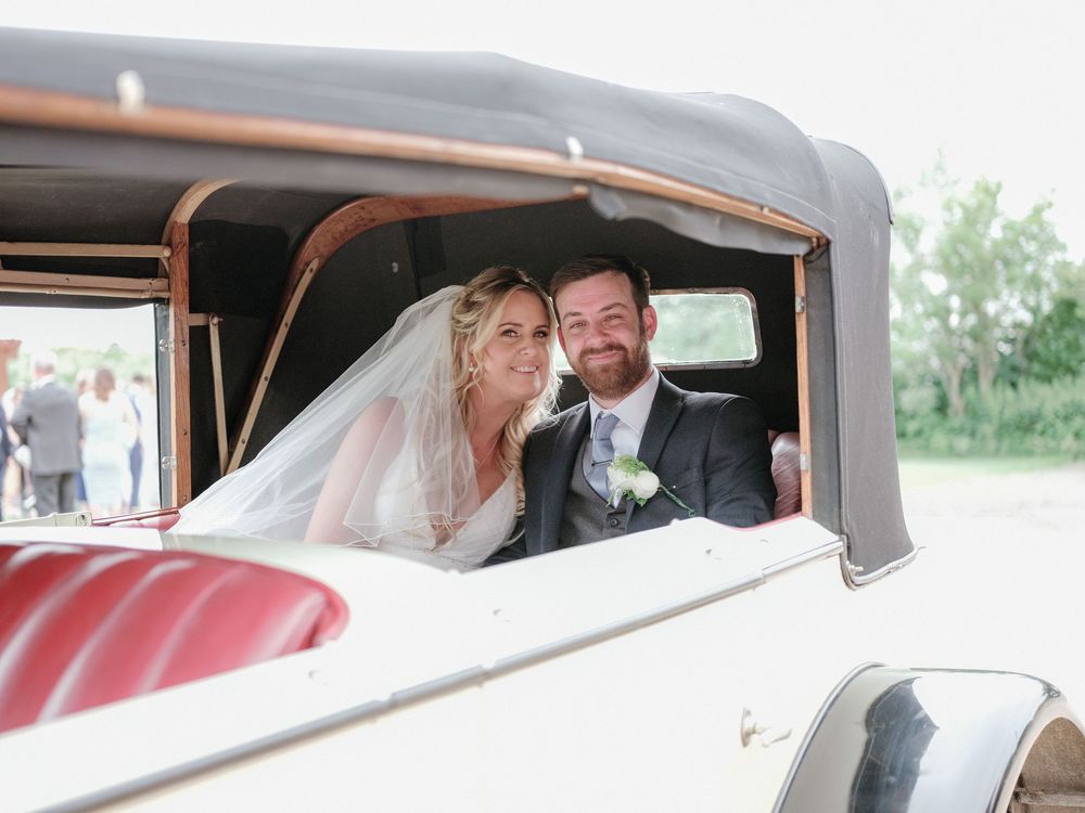A happy couple looks out of their vintage getaway car window after their summer wedding celebration.