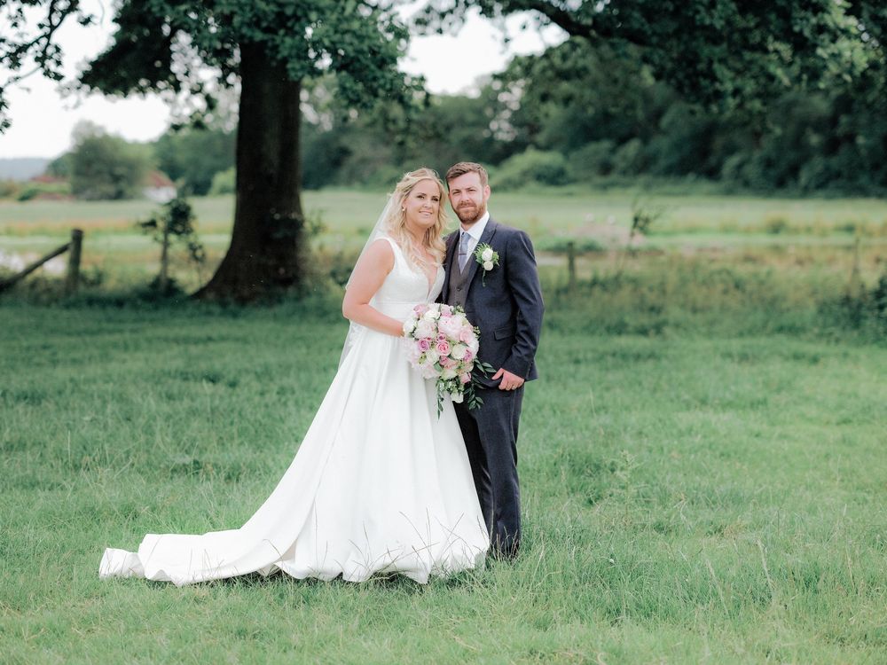A wedding couple stands together in a lush green field with a large tree in the background.