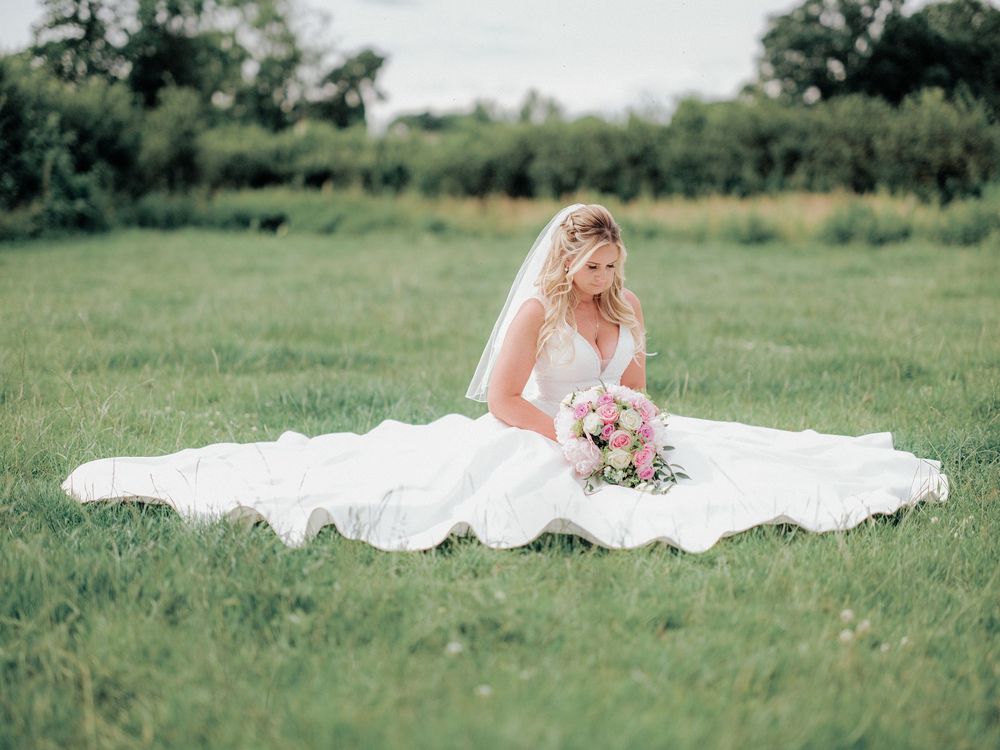 A bride in a flowing white gown sits gracefully in a green field while holding a pink and white bouquet.