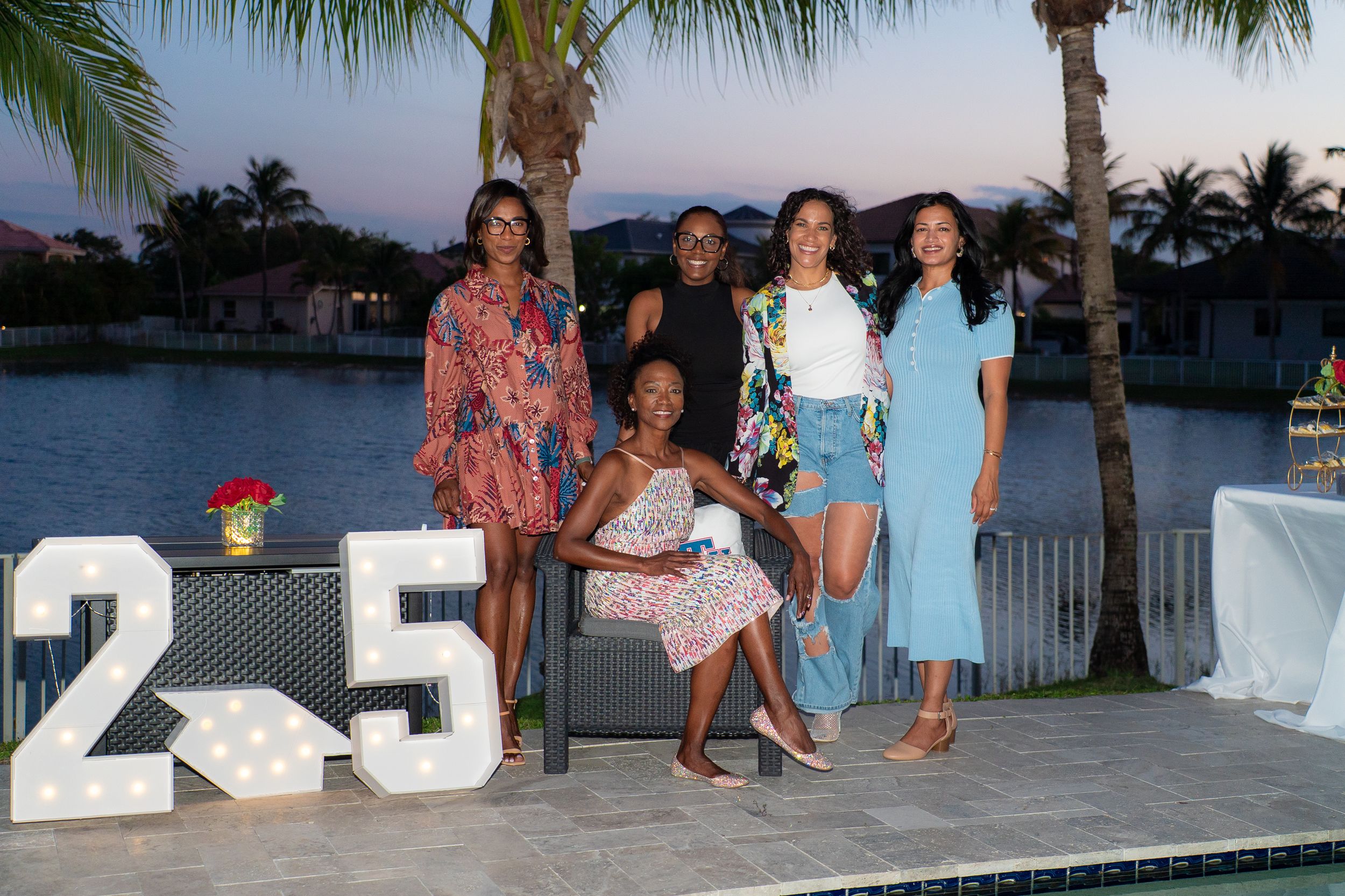 Group of five women posing at dusk by a pool with decorative number 25 marquee light-up letters.