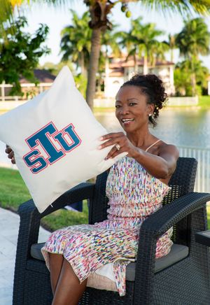 Woman smiling and holding an STU college pillow while seated outdoors by a tropical waterfront.