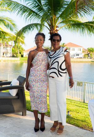 Two women smiling together outdoors at a tropical waterfront venue with palm trees in background.