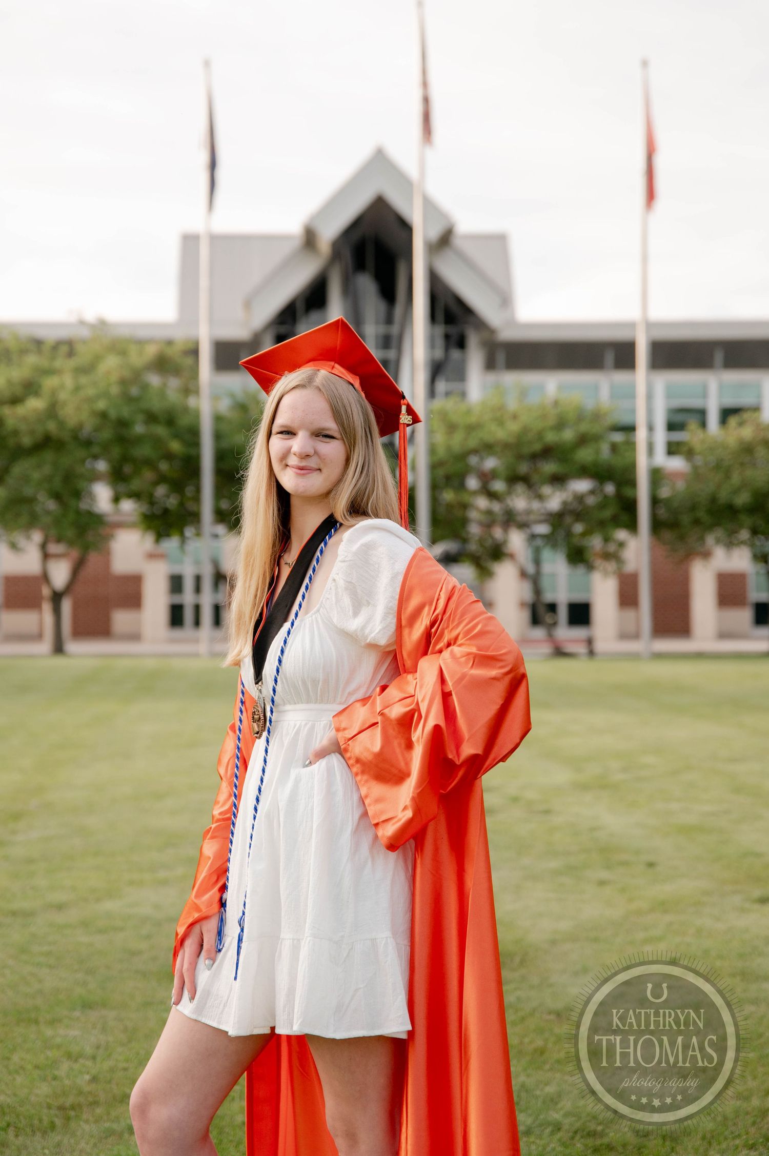 Graduate in red cap and gown stands on campus lawn with modern university building and flagpoles in background.
