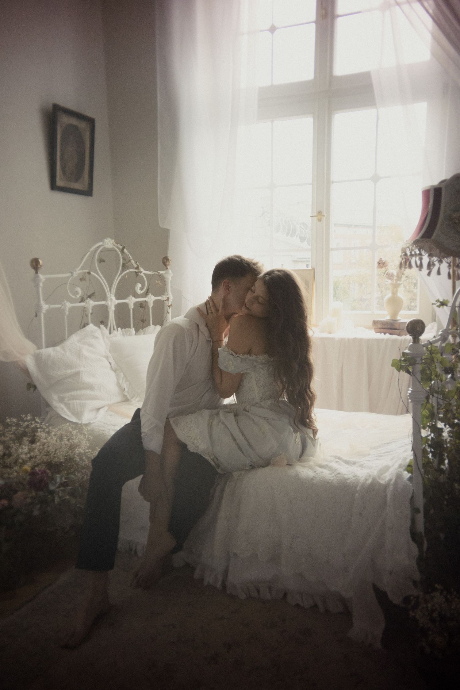 A romantic couple share an intimate moment in a sunlit bedroom with white decor and floral accents.