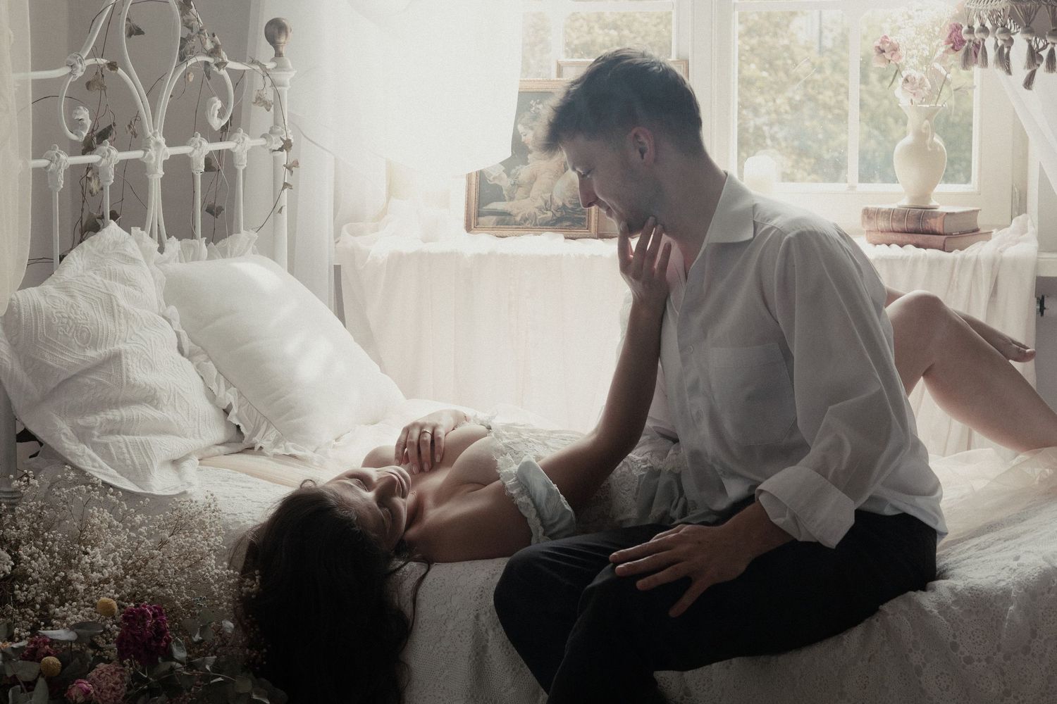 Two people share a tender moment together in a sunlit bedroom with white decor and floral elements.