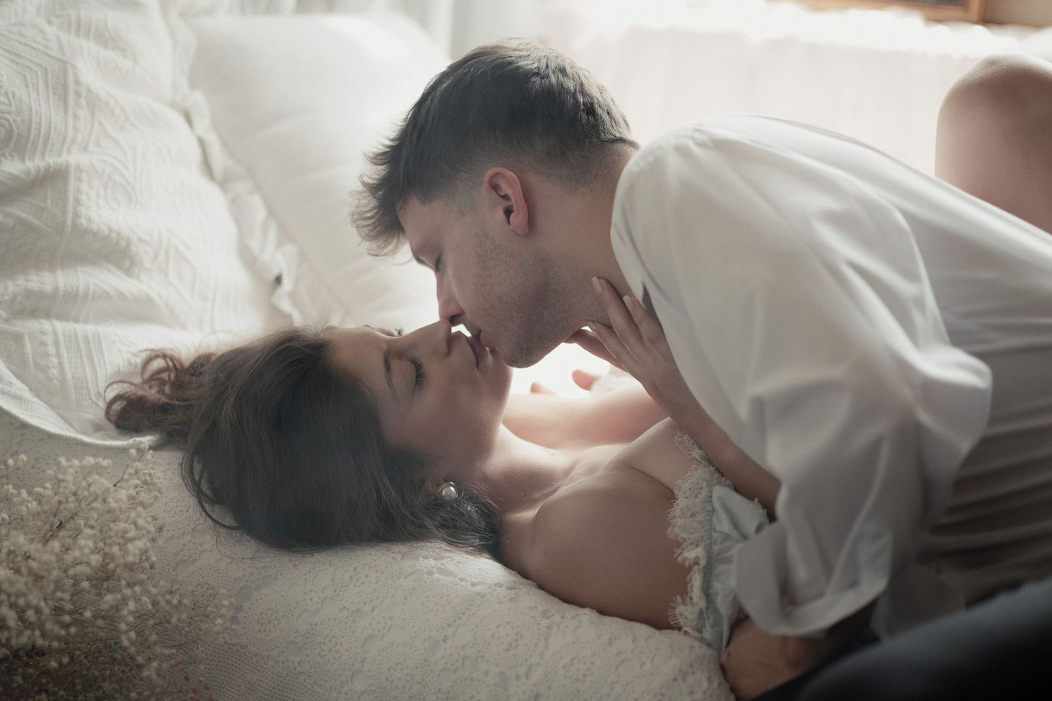 A couple shares an intimate romantic moment on a white bed with soft natural lighting.