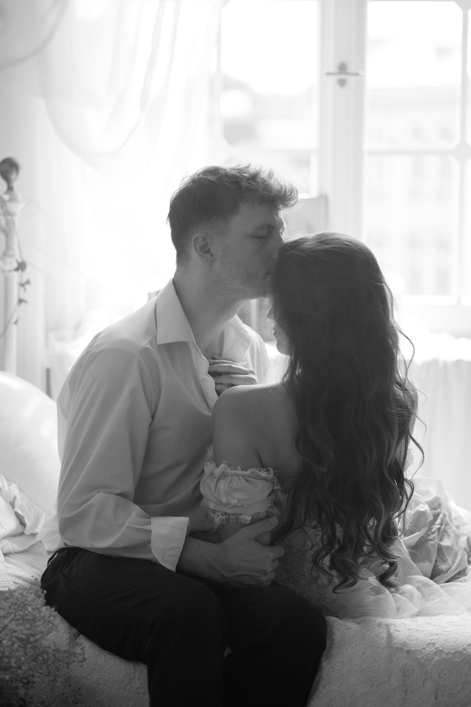 Intimate black and white photo of a romantic couple in an embrace by a window with soft natural lighting.