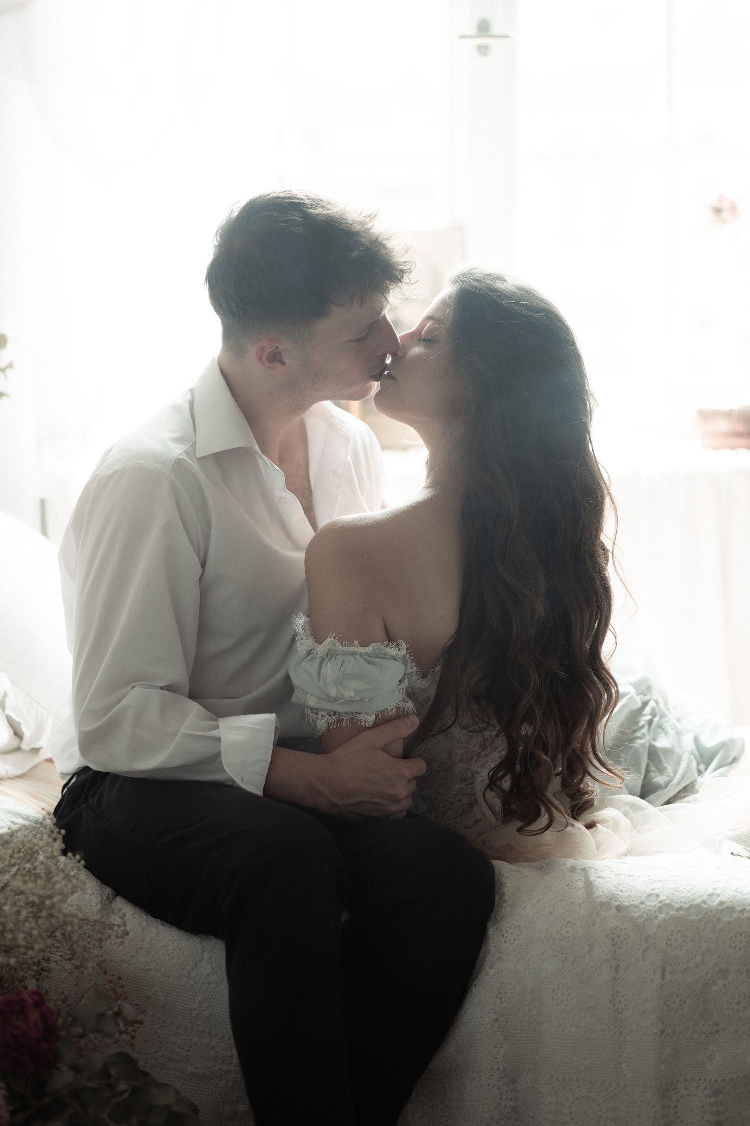 A romantic couple embraces in soft window light, sharing an intimate kiss against a white backdrop.