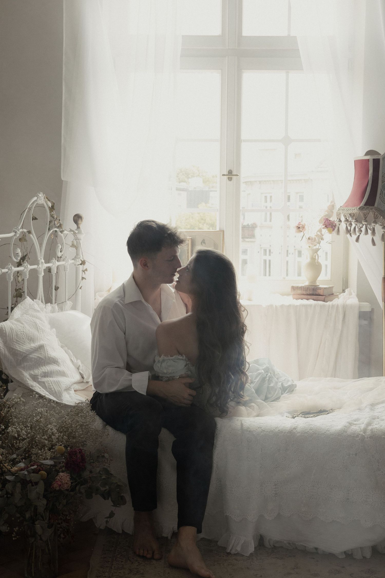 A romantic couple sits together in a dreamy sunlit bedroom with white decor and a vintage iron bed frame.