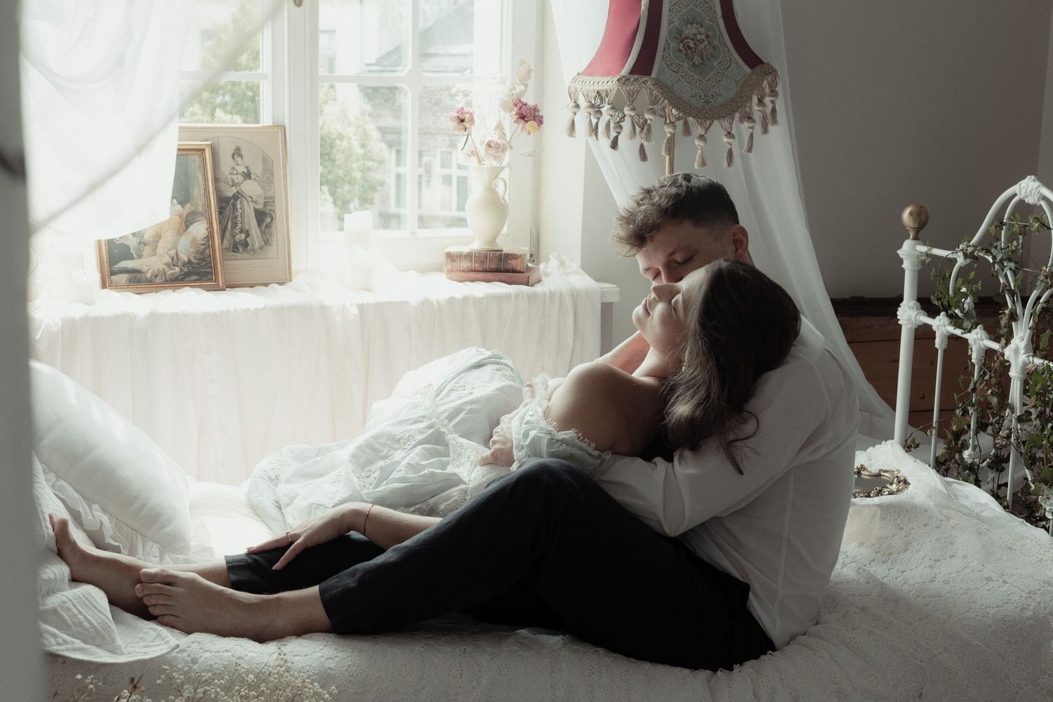 A person lies on white bedding while curled up against pillows near a sunny window.