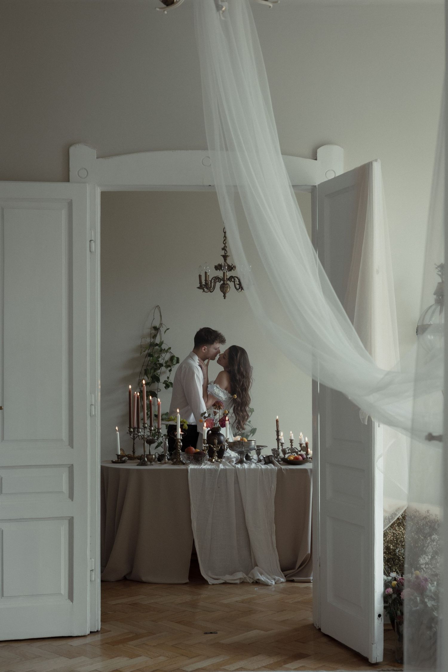 An elegant all-white wedding setup with flowing sheer drapes, double doors, and a table decorated with candelabras.