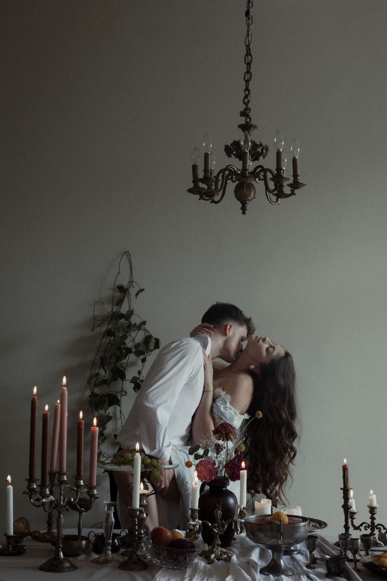 A romantic candlelit scene with antique chandelier, candlesticks, and flowers on a vintage table in a moody atmosphere.