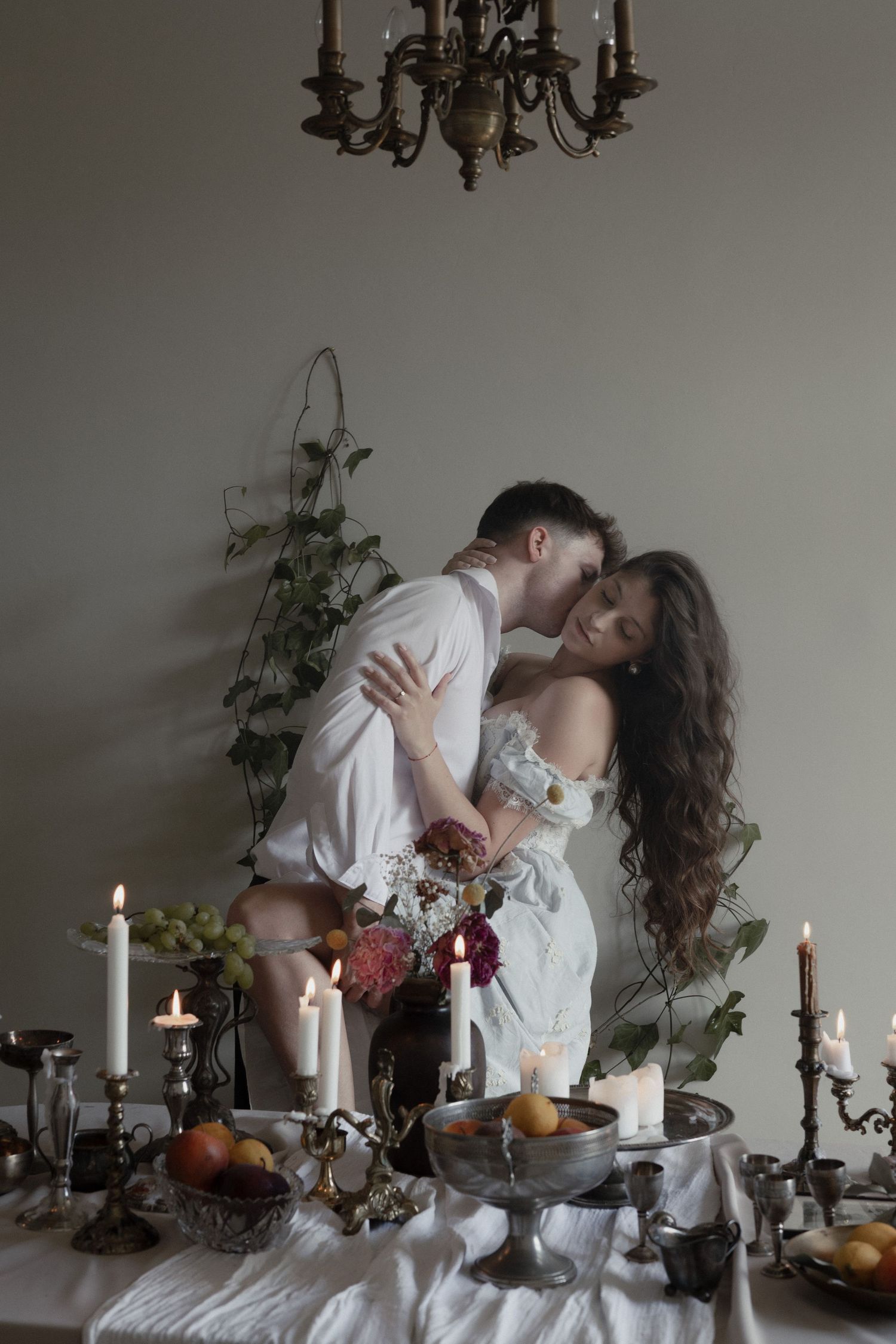 A romantic table setting with candles, fruits and greenery as a couple embraces in white clothing under a chandelier.