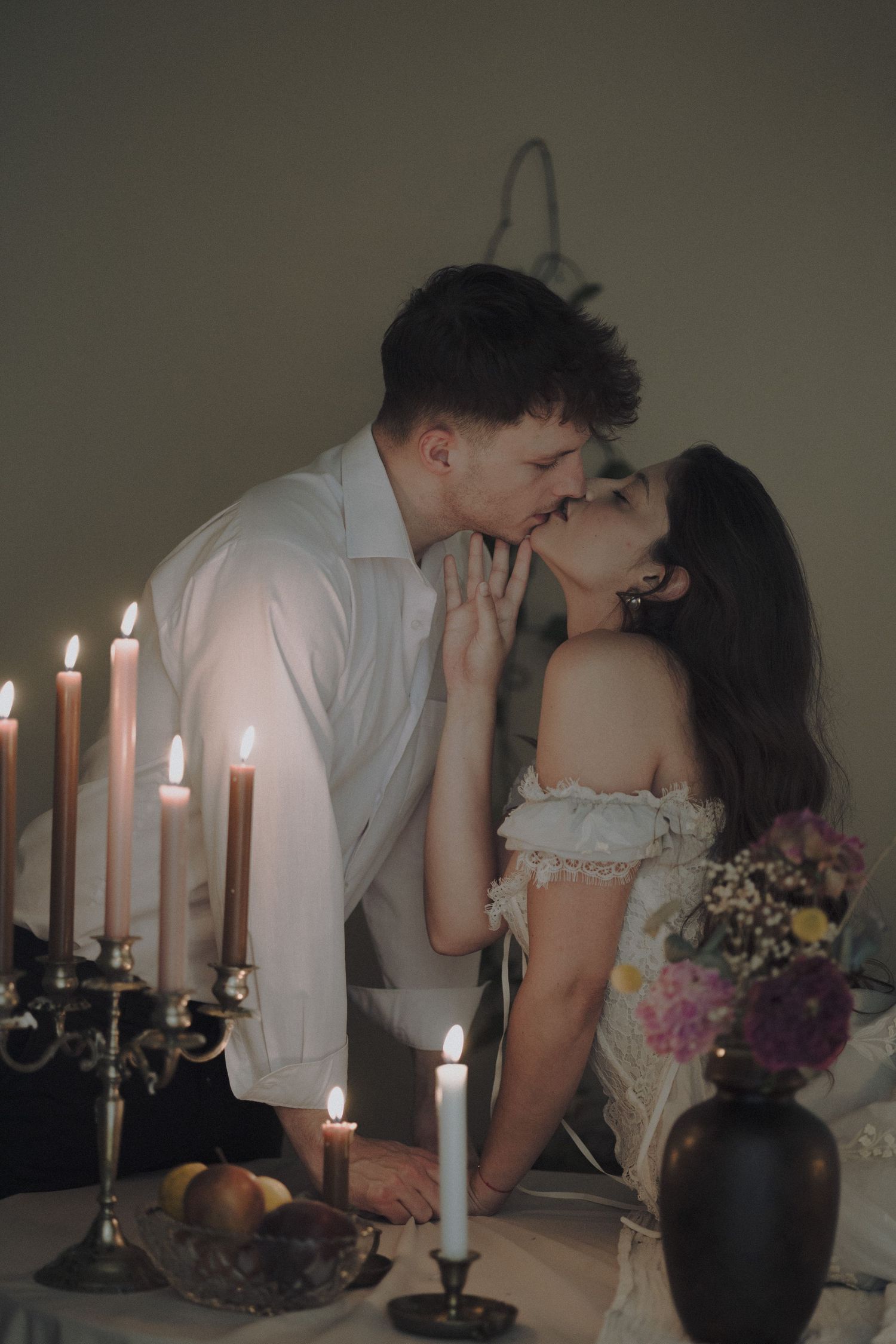 A romantic candlelit scene with a couple sharing an intimate moment alongside candelabras, flowers and fruit on a table.